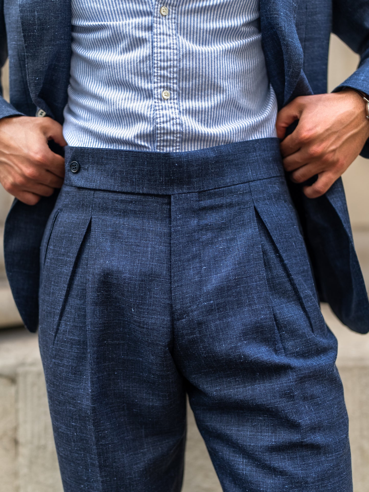 Hands hold the tailored light blue linen-wool trousers to highlight the belt-free waistline and functional side fasteners