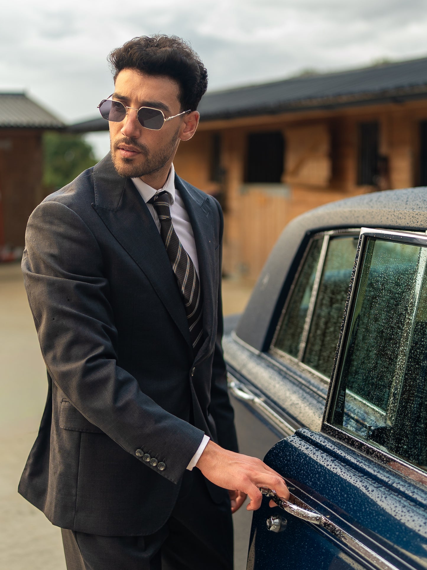A man in a tailored dark sharkskin grey blazer opens the door of a classic car, subtle sheen, structured shoulders, and quiet authority in motion.