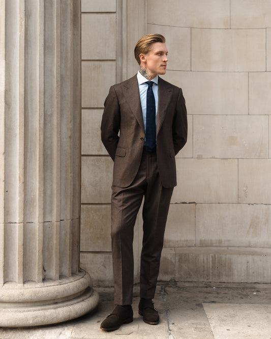 A man in a made-to-measure deep brown houndstooth suit stands with hands clasped behind his back.