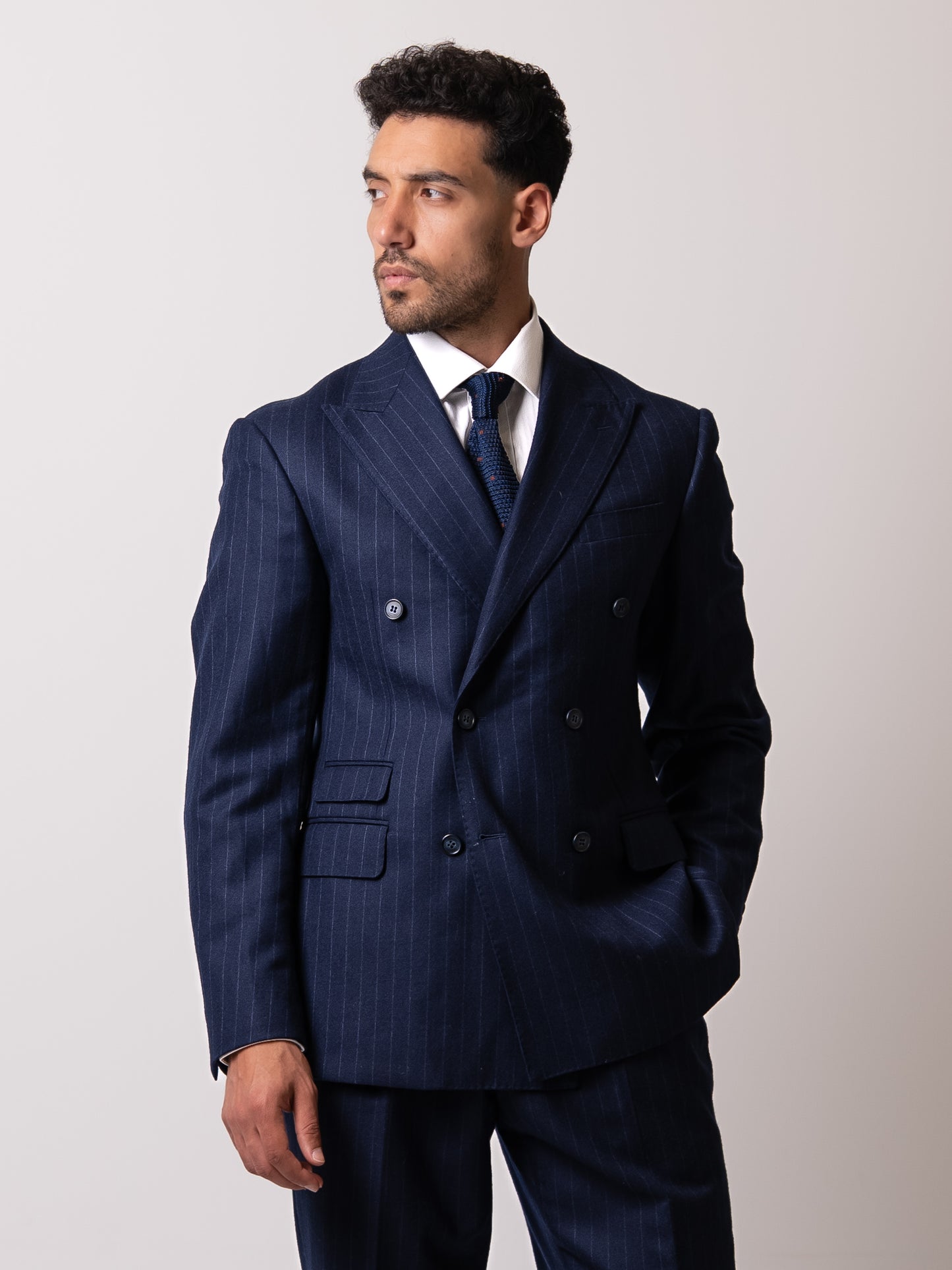 A man in a chalk stripe double-breasted blazer stands with quiet assurance, gaze turned left. Clean lines, commanding silhouette, and refined presence in natural light.