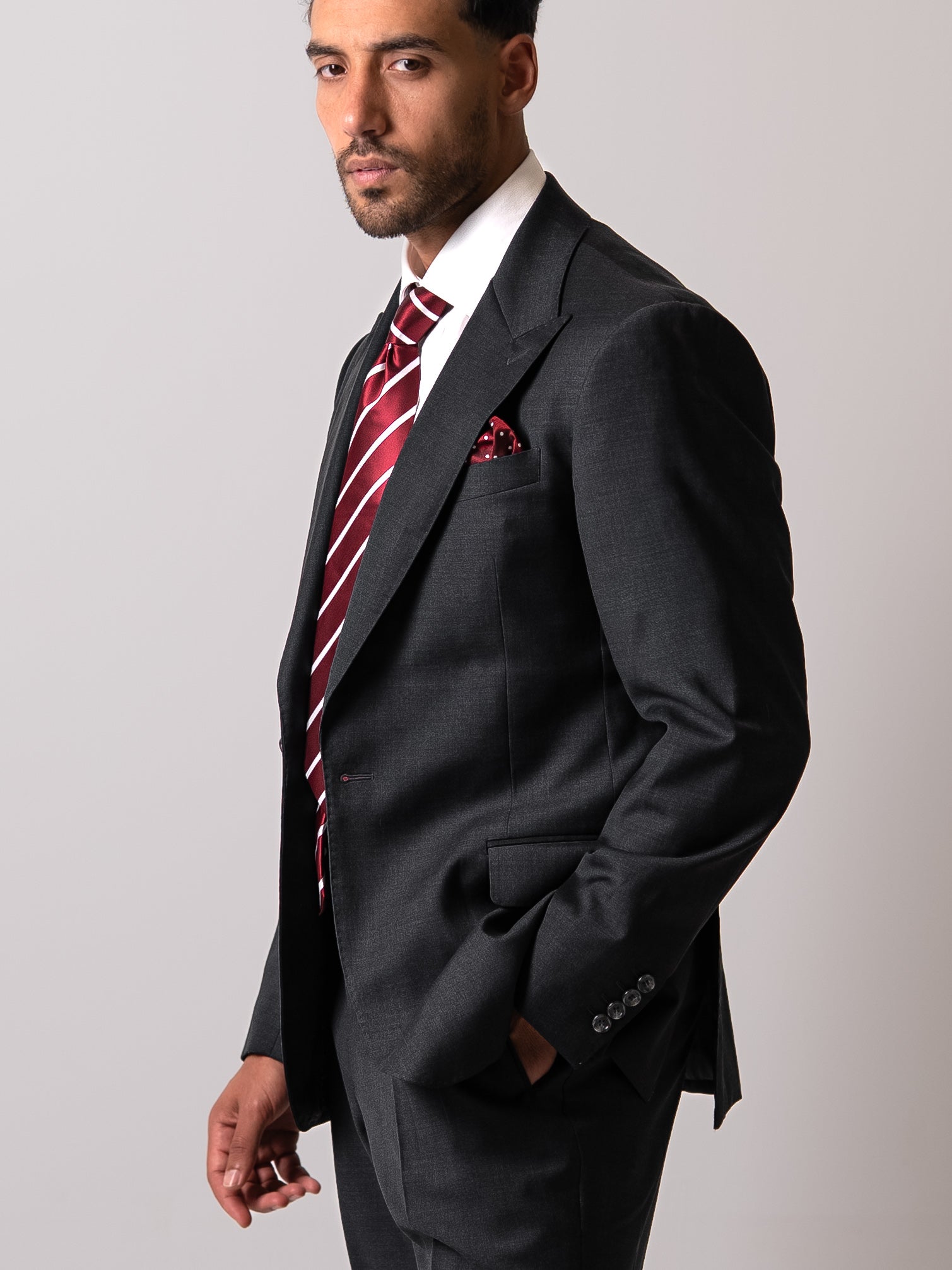 Side view of a man in a dark sharkskin grey blazer with hand resting in the flap pocket. Showcasing the blazer’s fluid drape, clean silhouette, and effortless ease of movement. 