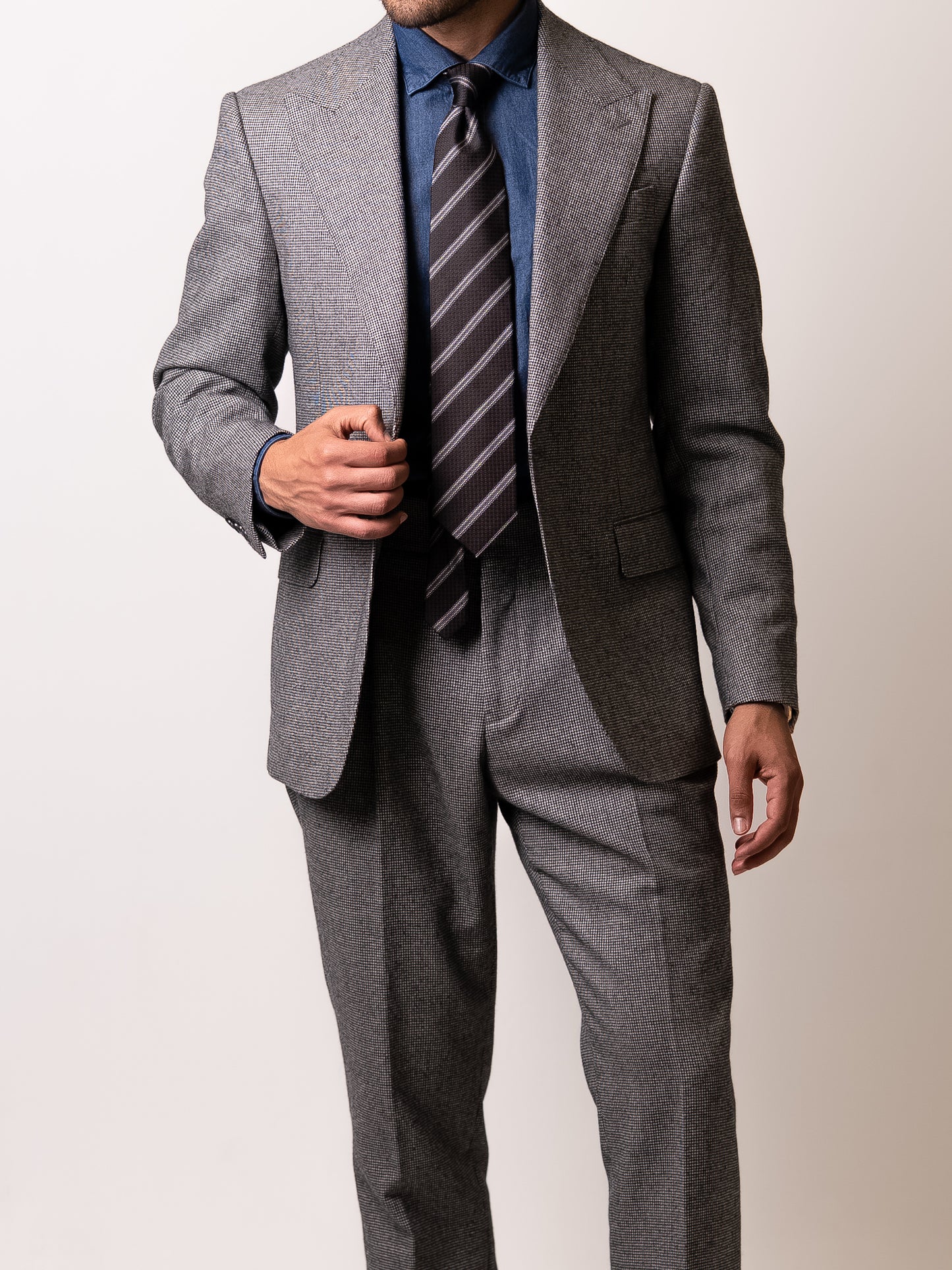 A man gently holds the lapel of his made-to-measure light grey houndstooth suit 
