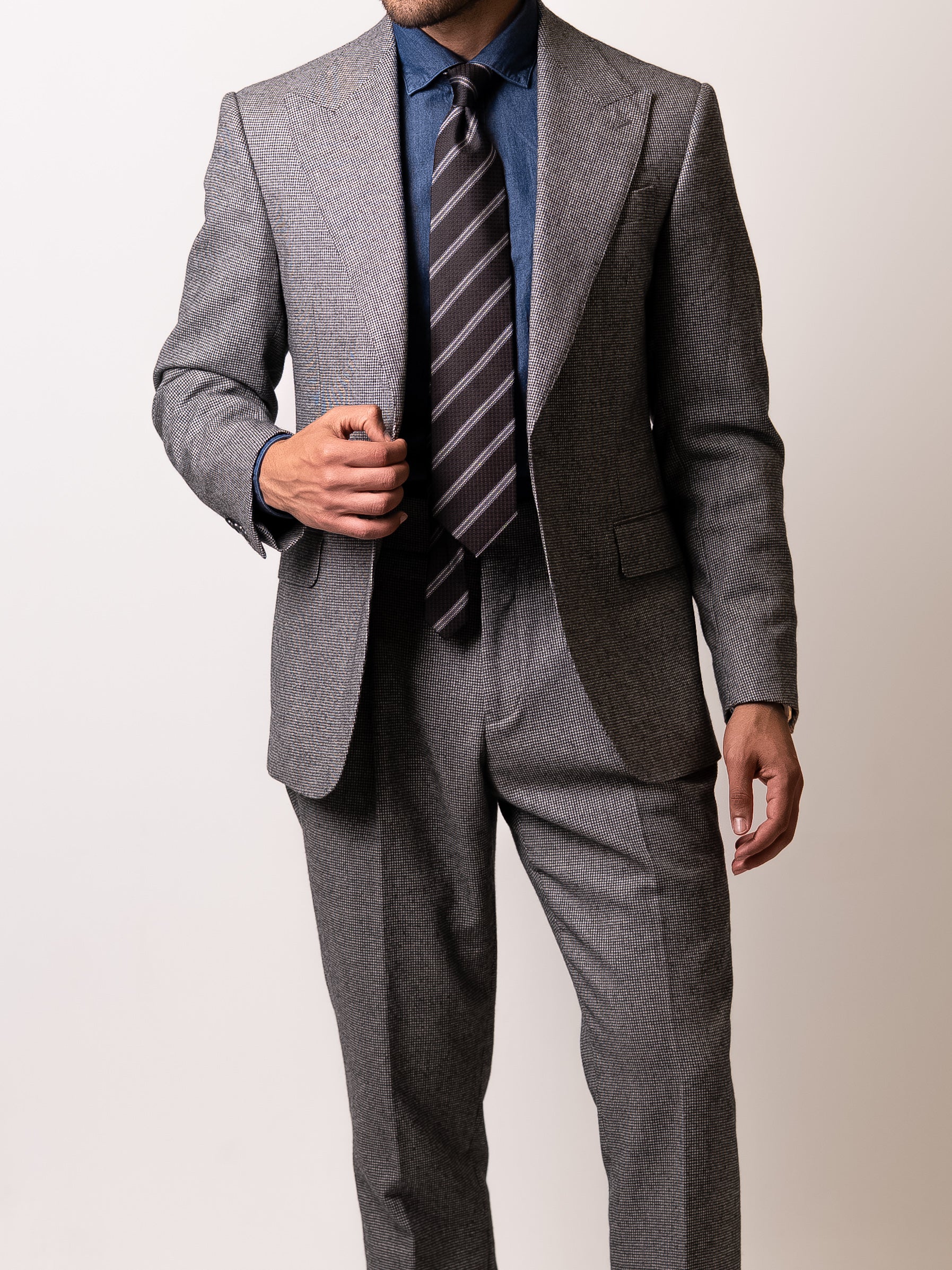 A man gently holds the lapel of his made-to-measure light grey houndstooth suit 
