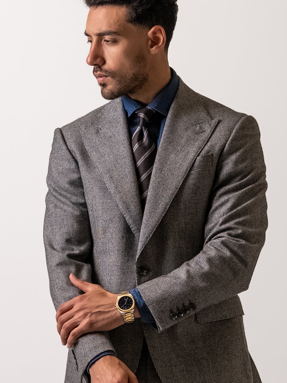 A man stands with his left hand resting on his right forearm in a tailored light grey houndstooth blazer
