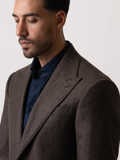 Close-up of the wide peak lapel on a deep brown houndstooth blazer — bold framing, textured weave, and sharp tailoring that defines presence without noise. 
