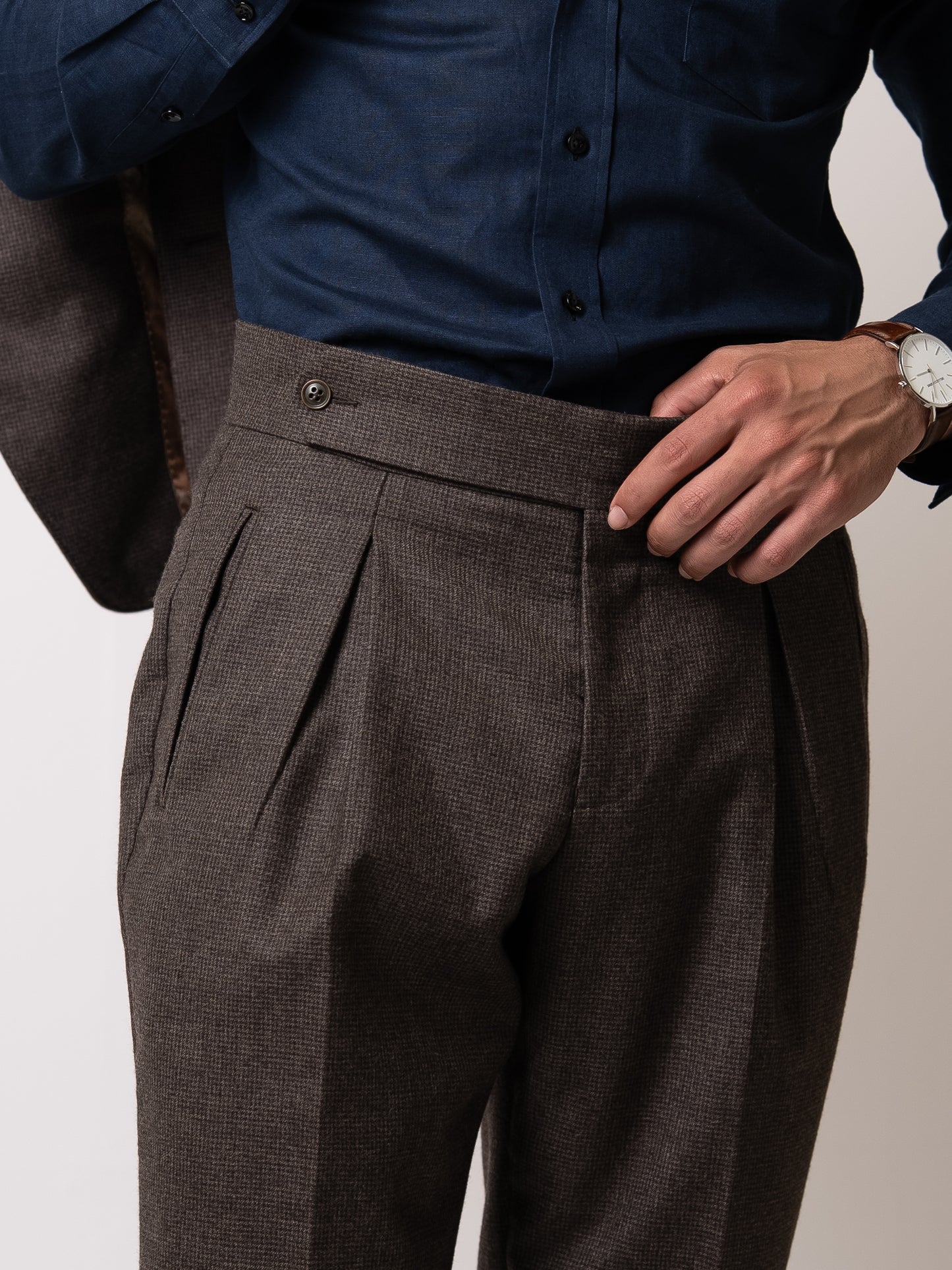 A man holds the front of his tailored deep brown houndstooth trousers to reveal the side fasteners 