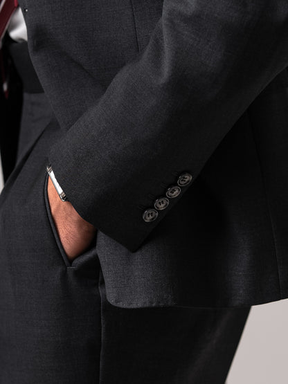 Side view of a man in a dark sharkskin grey suit with hand resting in the flap pocket. Showcasing the blazer’s fluid drape, clean silhouette, and effortless ease of movement. 
