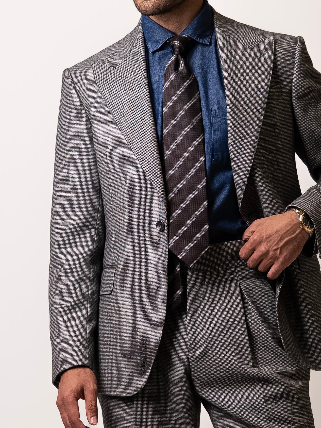 A man in a tailored light grey houndstooth blazer holds his trousers slightly to reveal the single-button closure