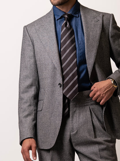 A man in a tailored light grey houndstooth blazer holds his trousers slightly to reveal the single-button closure