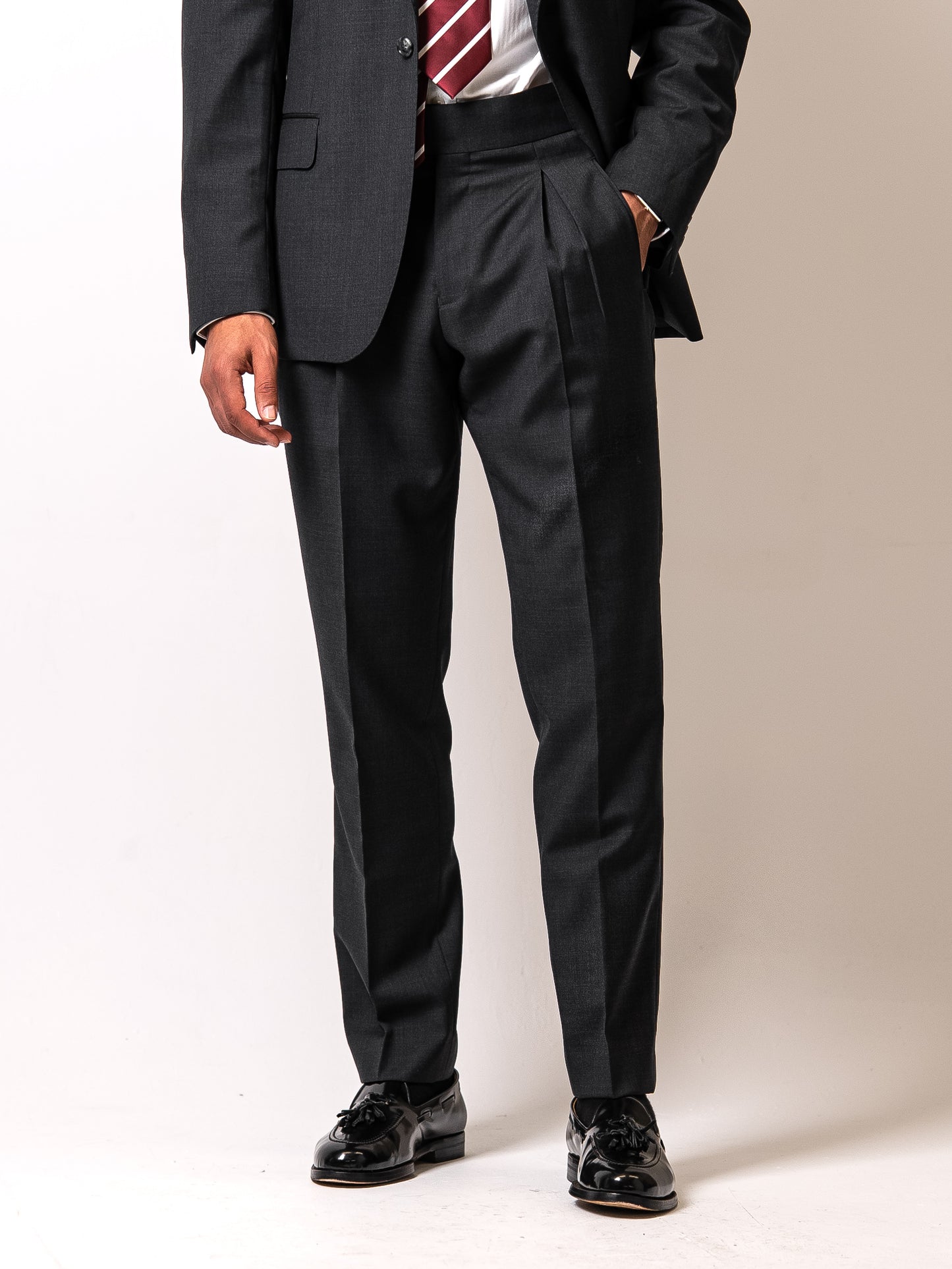 Front view of dark sharkskin trousers as the man rests his hand in the flap pocket — double pleats, side adjusters, and elegant drape in a refined stance.
