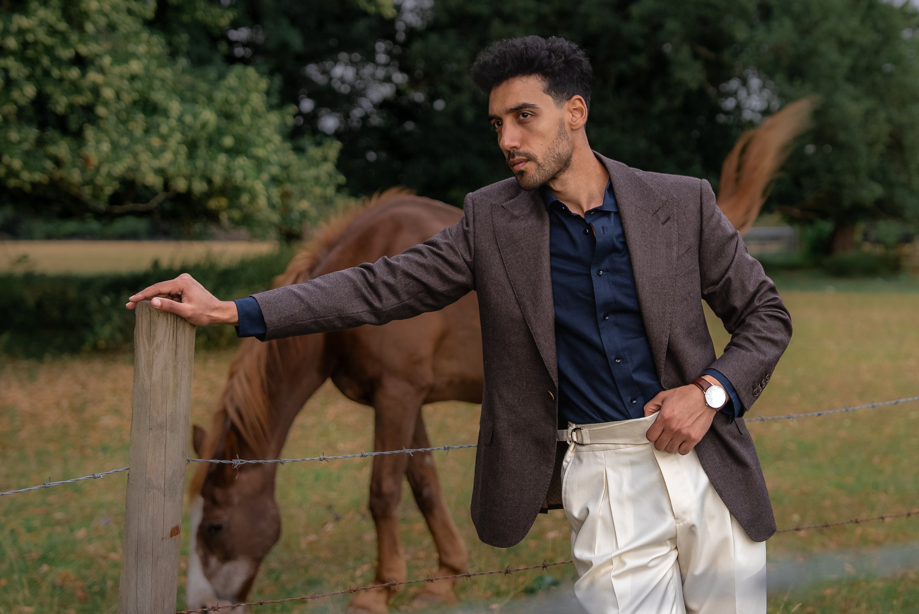 A man in a deep brown blazer and tailored white trousers poses with understated ease before a horse with earthy tones, refined contrast, and quiet strength
