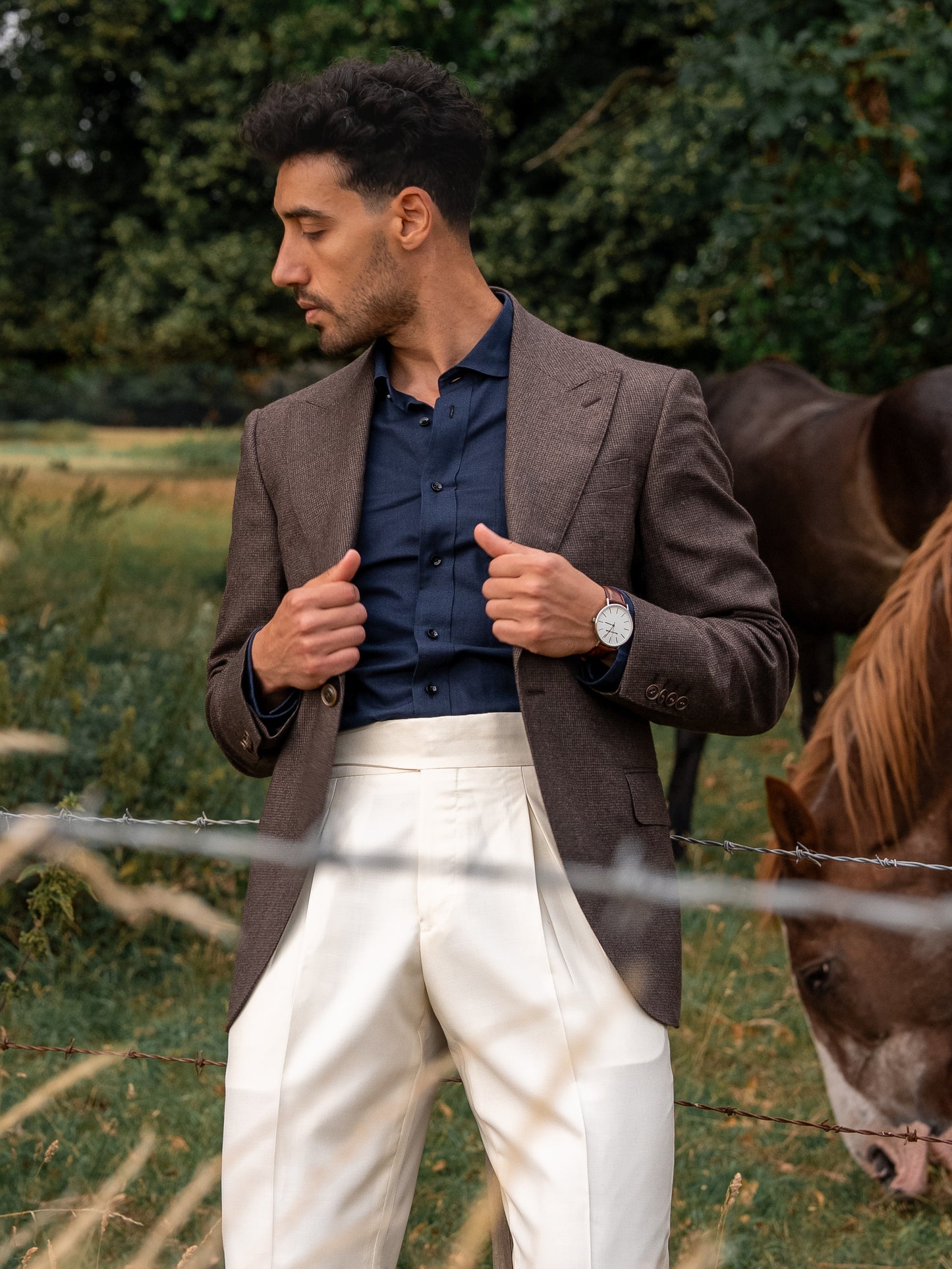 A man wearing a tailored white trousers worn with a deep brown houndstooth blazer, a horse standing nearby — earthy tones meet pure form in a moment of quiet strength and natural harmony. 
