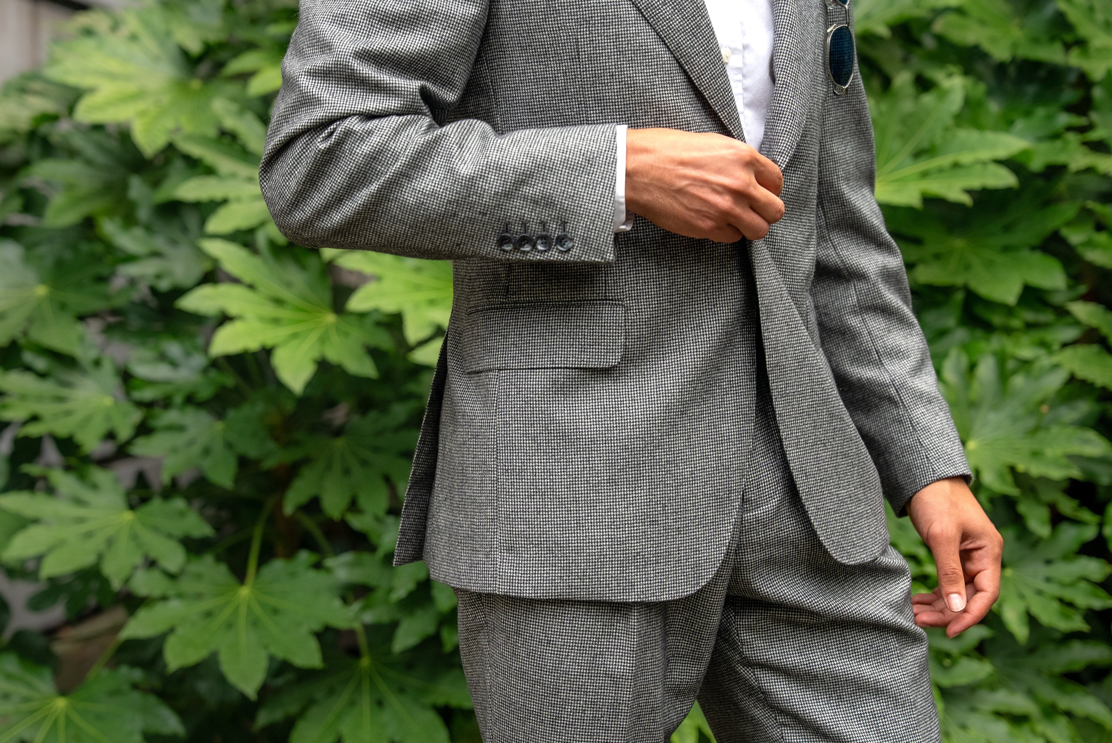 A man in a light grey houndstooth suit stands with poised confidence  structured shoulders, single-button closure, and elegant restraint in an urban setting. 
