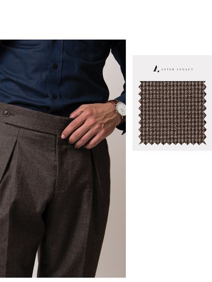 A man holds the front of his deep brown houndstooth trousers, revealing double pleats, side fasteners, and the rich wool fabric’s warm texture and subtle check