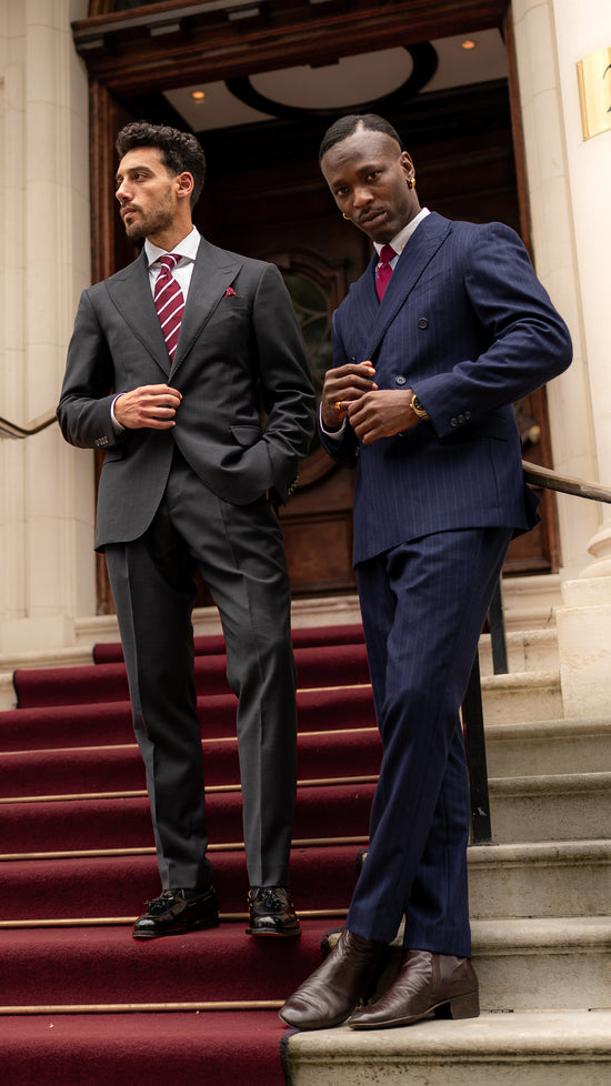 Two gentlemen in made-to-measure Aster Legacy suits: one in a dark grey sharkskin suit with subtle sheen and clean lines, the other in a chalk stripe double-breasted suit with wide peak lapels. Both embodying quiet authority, precision tailoring, and British craftsmanship. 
