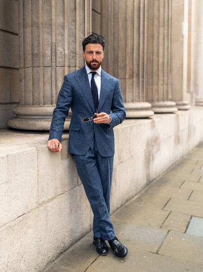 A man leans against a sunlit wall in made-to-measure a light blue linen-wool suit, sunglasses resting in his hand
