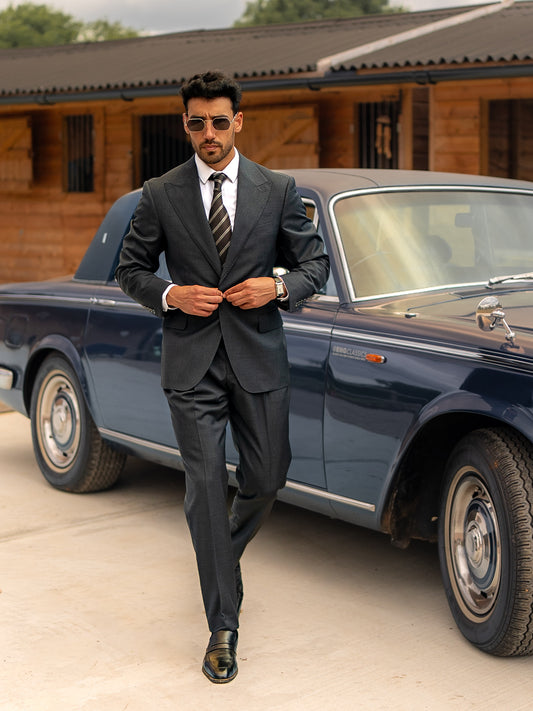A man in a made-to-measure dark sharkskin grey suit closes his single-button jacket while walking past a classic car