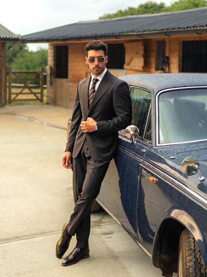 A man stands with legs crossed next to classic car in a made-to-measure dark sharkskin grey suit 