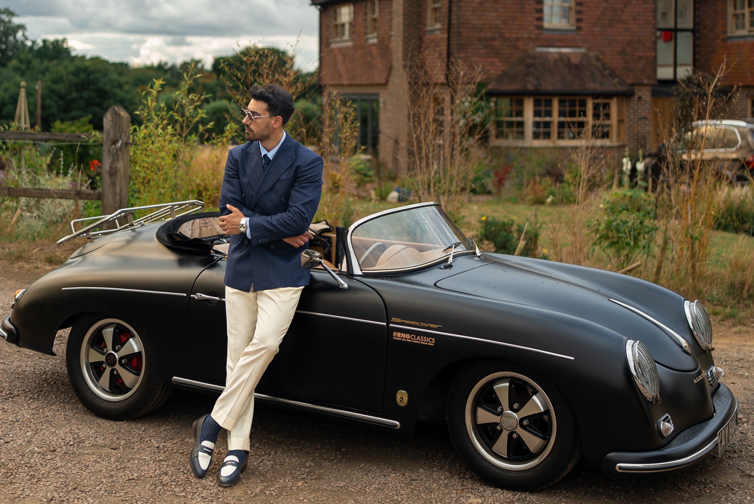 A man in a made-to-measure chalk stripe double-breasted blazer and tailored white trousers stands with quiet authority beside a vintage car 
