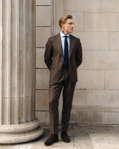 A man in a made-to-measure deep brown houndstooth suit stands with hands clasped behind his back.