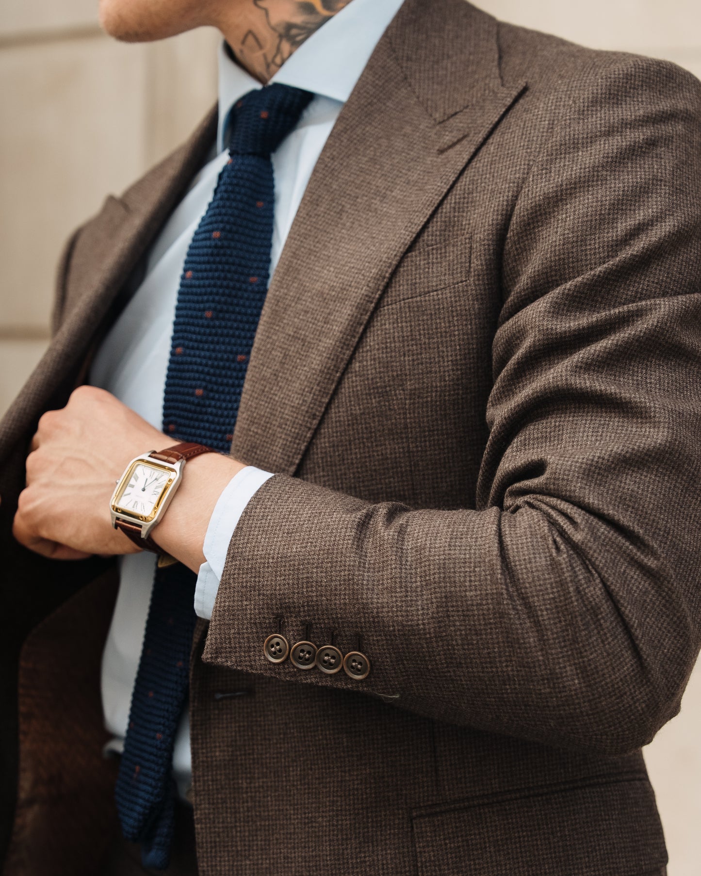 A man rests his hand inside the chest pocket of his made-to-measure deep brown houndstooth suit, a brown leather-strap watch visible.