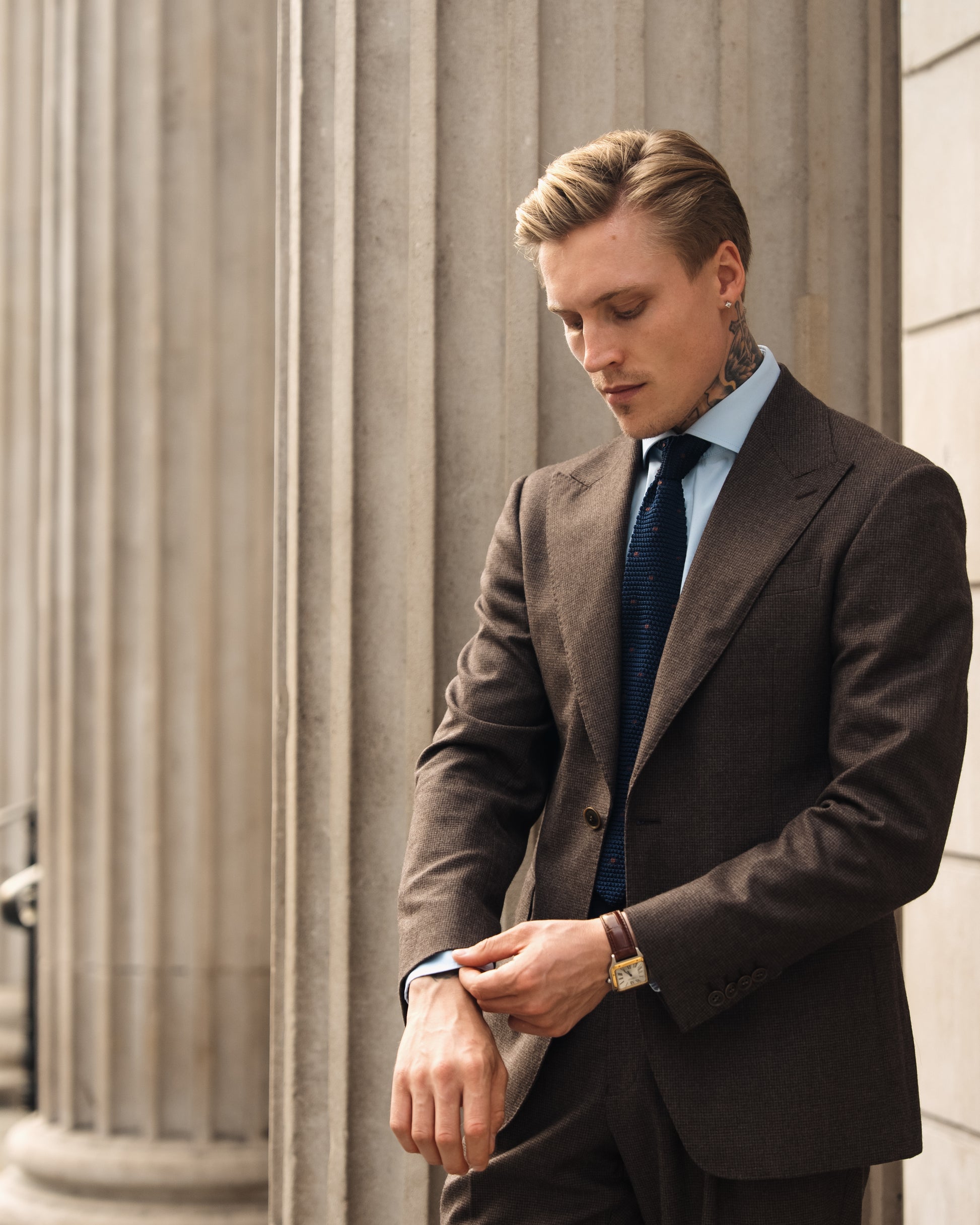 A man on a city street adjusts the cuff of his made-to-measure deep brown houndstooth suit, showcasing fluid drape, heritage texture, and effortless attention to detail.
