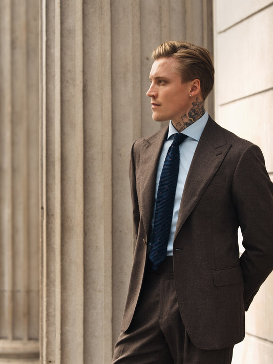A man stands in a tailored deep brown houndstooth blazer. Wide peak lapels, single-button closure, and structured shoulders conveying warmth, depth, and modern composure.